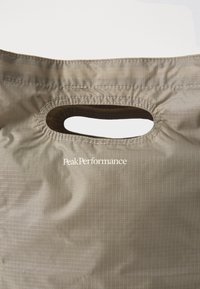 Heled pruun kangast kott ovaalse lõikega käepideme ja väikese valge "PeakPerformance" logo printimisega käepideme all.