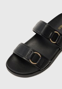 Svarta sandaler med två breda remmar som har guld ringdetaljer. Slät läderyttextur, platt sula och minimalistisk design.