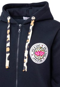 Schwarze Zip-Hoodie mit Leopardenmuster bei den Kordeln und einem runden Grafik-Patch, das Herzen und den Text "wild at heart" zeigt. Weicher Stoff.