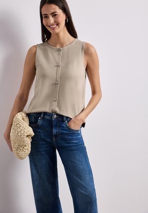 Smilende kvinde iført beige ærmeløs knapbluse, blå jeans, holder en beige flettet clutch, hånd i lommen, mod en lys baggrund.