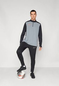 Nike-Sportbekleidungsset mit einem grauen Langarm-Pullover mit schwarzen Akzenten und einem halben Reißverschluss, kombiniert mit schwarzen, zulaufenden Hosen und einem Fußball.
