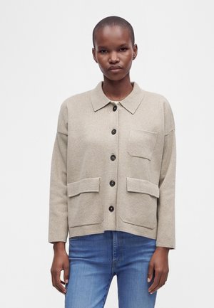 Femme portant une veste beige à boutons avec trois poches avant et un jean bleu, debout devant un fond blanc uni.