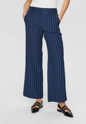 Pantalones de pierna ancha azul marino con rayas verticales blancas, combinados con zapatos planos negros puntiagudos con hebillas plateadas.