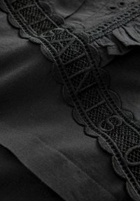 Tela negra con intrincados detalles de encaje negro, que presenta un borde festoneado y patrones geométricos, junto con acentos con volantes.
