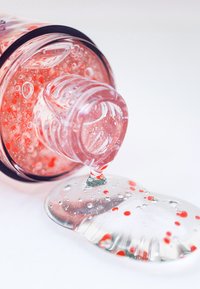 Transparent flaske med en pink gel, der strømmer ud, indeholdende røde perler. Glat tekstur, blank finish og bobler synlige i gelen.