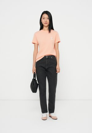Perzikkleurig t-shirt met korte mouwen en logo, gecombineerd met hoge zwarte jeans. Het model houdt een kleine zwarte tas vast en draagt lichtroze flats.