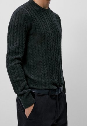 Uomo che indossa un maglione verde scuro a trecce con le mani in tasca, abbinato a pantaloni neri e una cintura nera su uno sfondo semplice.