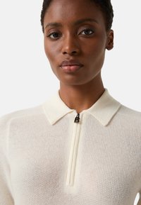 Kremowy sweter z dzianiny z kołnierzem na suwak. Charakteryzuje się fakturowaną tkaniną i subtelnym wzorem. Czarna zawieszka na suwaku z literą "B".