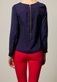 Blusa de seda azul marino con mangas largas y detalle de cremallera en la espalda, combinada con vaqueros ajustados de color rojo brillante. Textura suave, diseño entallado.