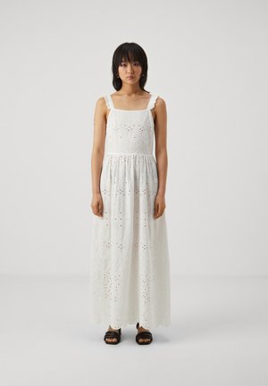 Robe maxi blanche avec des broderies complexes et des motifs à œillets, dotée de fines bretelles et d'une taille cintrée. Portée avec des sandales noires.
