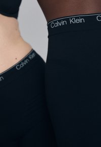 Mustad venivast kangast retuusid Calvin Kleini logoga vöökohaga, millel on sile tekstuur ja keha järgiv lõige, kujutatud kahel figuuril.