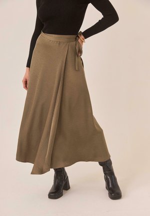 Mujer con falda larga cruzada de color oliva, top de manga larga negro con textura acanalada, botines negros con tacón y una pulsera en la muñeca derecha.