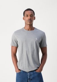 Grå bomulds-T-shirt med korte ærmer, rund halsudskæring og et CK-logo på venstre bryst. Båret med blå denimjeans.