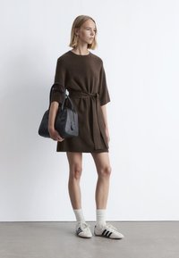 Braunes Strickkleid mit kurzen Ärmeln und einem Gürtel, kombiniert mit weißen Sportschuhen und grauen Socken, dabei eine schwarze Handtasche haltend.