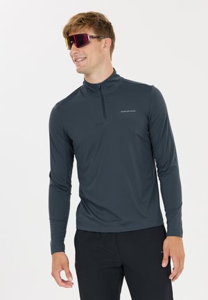 Man glimlacht, draagt een donkergrijze lange mouwen sporttop met een kwartrits en gespiegelde sportzonnebril.