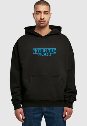 Schwarzer Hoodie mit dem Text "NOT IN THE MOOD" in Blau über der Brust, Fronttasche sowie gerippten Bündchen und Saum. Getragen mit blauen Jeans.