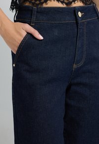 Pantaloni in denim blu scuro con vita alta, caratterizzati da cuciture gialle, una tasca frontale e chiusura a bottone metallico. Superficie strutturata visibile.