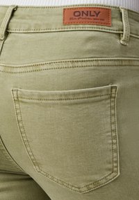 Olivno zelena denim tkanina z gladko teksturo, ki ima zadnji žep in rjavo usnjeno oznako z oznako "ONLY" vzorčeno na pasu.