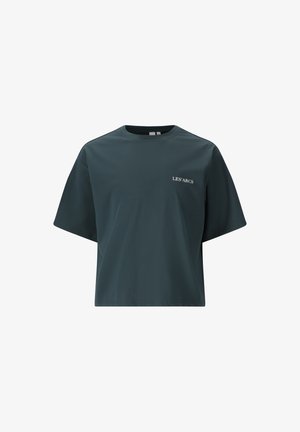 Jonge vrouw die loopt, gekleed in een donkergroen cropped T-shirt met de tekst "LES ARCS", zwarte wijde broek en zwarte sandalen tegen een witte achtergrond.