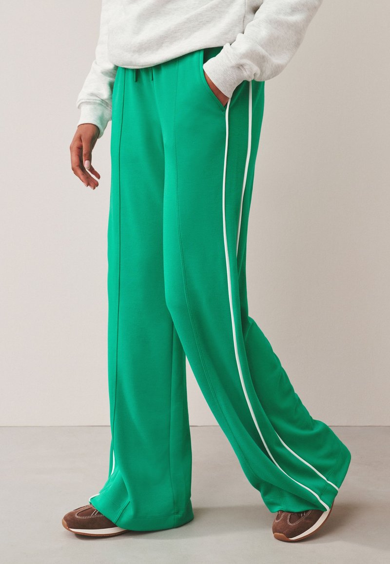 Personne portant un pantalon vert à jambes larges avec des bandes blanches sur les côtés, une main dans la poche, associé à des baskets marron et un sweat gris clair.