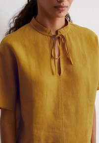 Mustard yellow short-sleeved top in lightweight fabric, featuring a band collar and a front keyhole with a tie closure.
