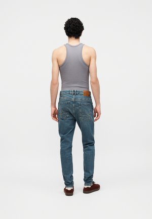 Mann mit lockigem Haar, trägt ein graues Tanktop, blaue Jeans und braune Schuhe, steht mit dem Rücken zu einem schlichten weißen Hintergrund.