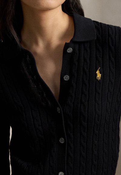 Close-up af en person iført en sort kabelstrikket cardigan med krave, knapper og en lille gul broderet hestelogo på brystet.