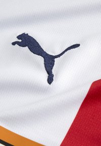 Hvidt stof med en fin tekstur, som har et broderet puma-logo i marineblå. Røde og orange detaljer langs den nederste kant.