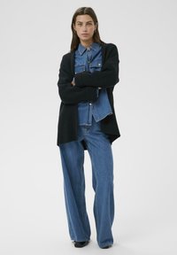 Frau mit verschränkten Armen, die ein blaues Jeanshemd und Jeans sowie einen schwarzen oversized Cardigan trägt, steht vor einem weißen Hintergrund.