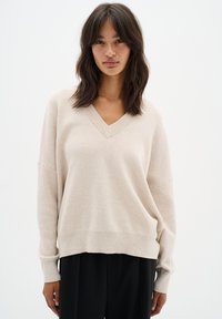 Sweater beige à col en V avec des manches longues et des épaulettes tombantes. Texture douce, coupe décontractée, bord inférieur au niveau des hanches. Associé à un pantalon noir.