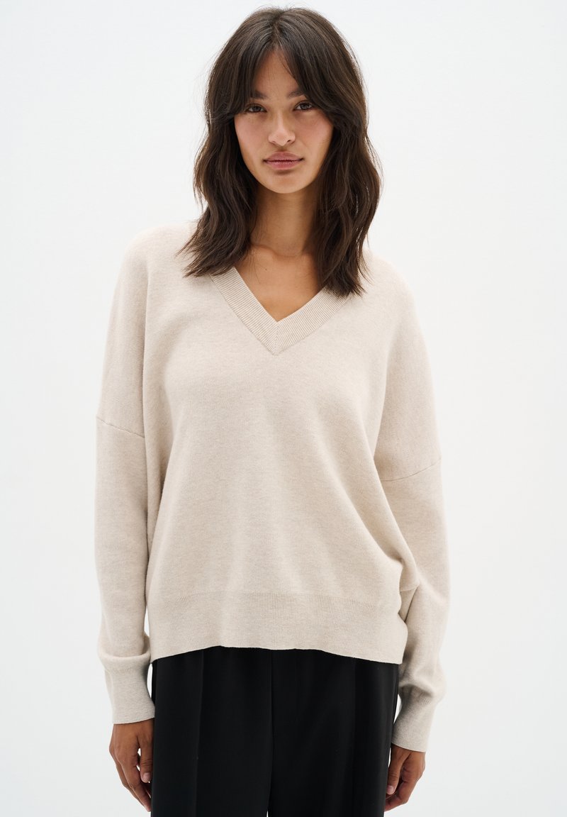 Sweater beige à col en V avec des manches longues et des épaulettes tombantes. Texture douce, coupe décontractée, bord inférieur au niveau des hanches. Associé à un pantalon noir.