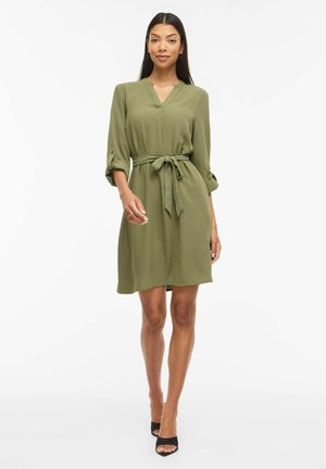 Femme debout portant une robe olive verte mi-longue à manches trois-quarts avec une ceinture nouée à la taille, associée à des talons ouverts noirs.