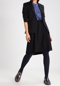 Svart kavaj över en blå knapplång skjorta, i kombination med en svart kjol och mörka strumpbyxor. Glansiga svarta skor med fransdetalj.