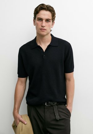 Giovane uomo con capelli corti castano chiaro che indossa una polo nera texturizzata a maniche corte e pantaloni scuri, tenendo una giacca color beige in una mano.