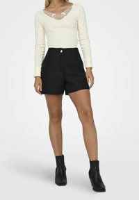 Cremfarbener Langarmoberteil mit Spitze am Ausschnitt, kombiniert mit hochgeschnittener schwarzer gemusterter Shorts. Das Model trägt schwarze Knöchelstiefel.