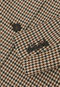 Brunt, sort og kremfarvet stof med hundetandsmønster og sorte knapper syet på overlappende klapper på ærmet af en skræddersyet jakke.