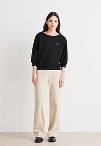 Felpa nera a maglia con scollo ampio e maniche a sbuffo, caratterizzata da un logo rosso ricamato. Abbinata a pantaloni chiari beige a vita alta.