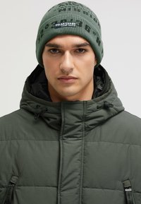 Green quilted jacket with a high collar and drawstring feature. A green knit beanie with black lettering sits on the head.