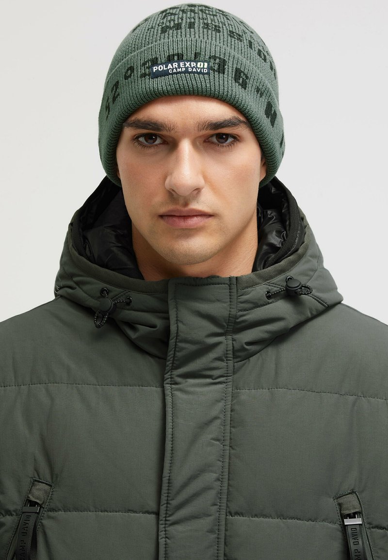 Green quilted jacket with a high collar and drawstring feature. A green knit beanie with black lettering sits on the head.