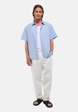 Homme debout portant une chemise à manches courtes rayée bleu clair sur un t-shirt blanc, un pantalon blanc ample et des sandales noires à brides.