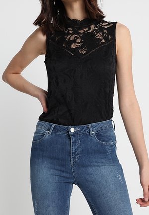 Vrouw draagt een mouwloze zwarte kanten top met bloemmotief en hoge halslijn, gecombineerd met blauwe high-waisted jeans.