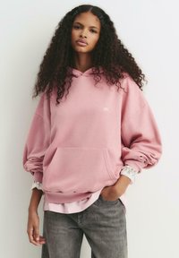 Ung kvinna med långt lockigt hår iklädd en lös rosa hoodie över en blus med spetsärmar och grå jeans, stående med en hand i fickan.