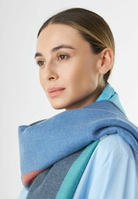 Écharpe en laine drapée autour du cou, présentant un mélange de rayures bleues, turquoise, grises et corail ; texture douce et design superposé.