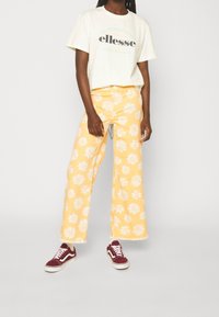 Gelbe weit geschnittene Hosen mit weißem Blumenmuster, kombiniert mit einem hellgrünen T-Shirt mit "ellesse"-Logo und kastanienbraunen Sneakers.