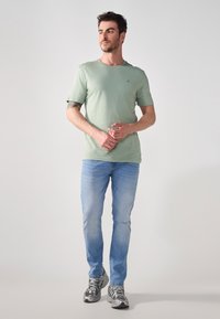 Világos zöld pamut póló rövid ujjakkal, világoskék farmernadrággal párosítva. Szürke sportcipő ezüst részletekkel egészíti ki az öltözetet.