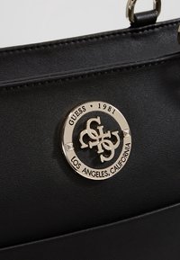 Czarna torebka z sztucznej skóry z okrągłym metalowym logo z wygrawerowanym napisem "GUESS 1981" i "LOS ANGELES, CALIFORNIA". Gładka tekstura, sztywny kształt.