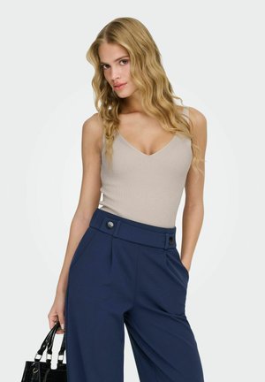 Jeune femme aux longs cheveux blonds ondulés portant un haut beige sans manches et un pantalon bleu marine taille haute, tenant un sac à main noir.