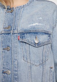 Světle modrá džínová bunda s klasickým límcem, kovovými knoflíky a předními kapsami s viditelným šitím a opotřebovanými detaily.