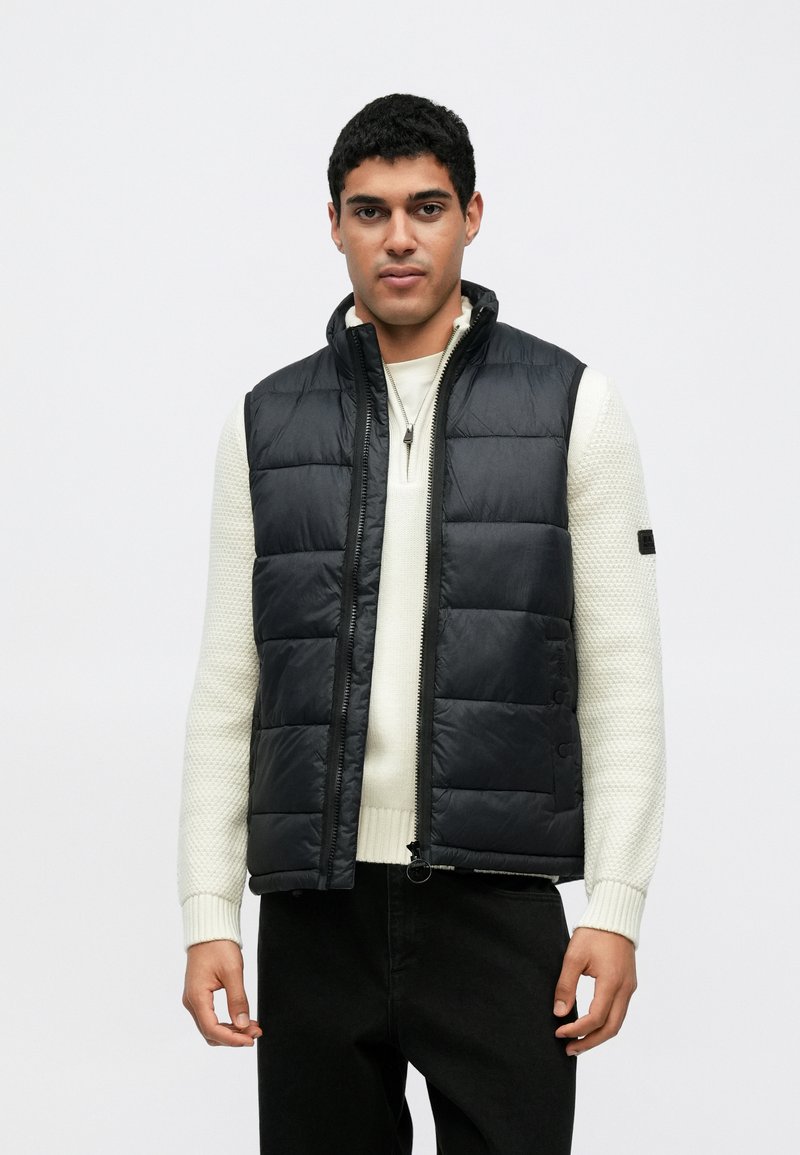 Μαύρο καπιτονέ γιλέκο με ψηλό γιακά, εμπρός φερμουάρ και πλαϊνά κουμπιά· φορεμένο πάνω από ένα μαλακό κρεμ πουλόβερ με υφή. Τοποθετημένο σε λευκό φόντο.