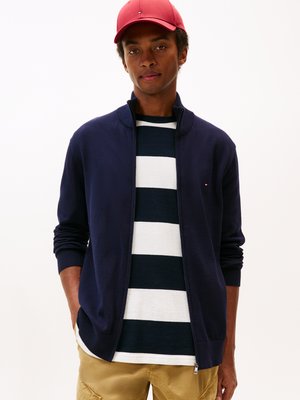 Giacca a zip blu navy con polsini a costine sopra una maglietta a righe blu navy e bianca. Il modello indossa un cappellino rosso, con le mani nelle tasche, e pantaloni khaki.
